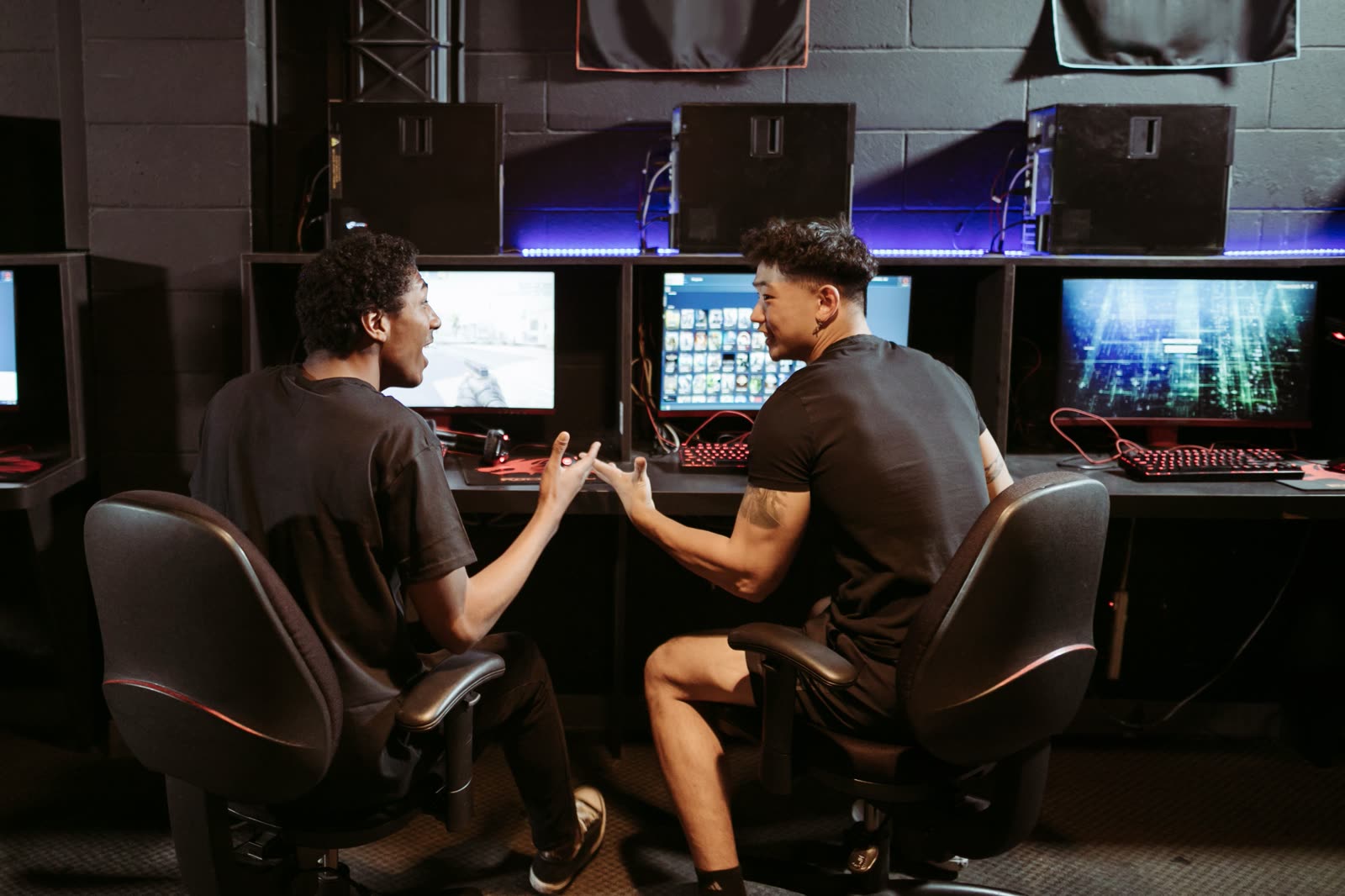 A long desk row of gaming stations with glowing monitors and black chairs at the NOPING computer club